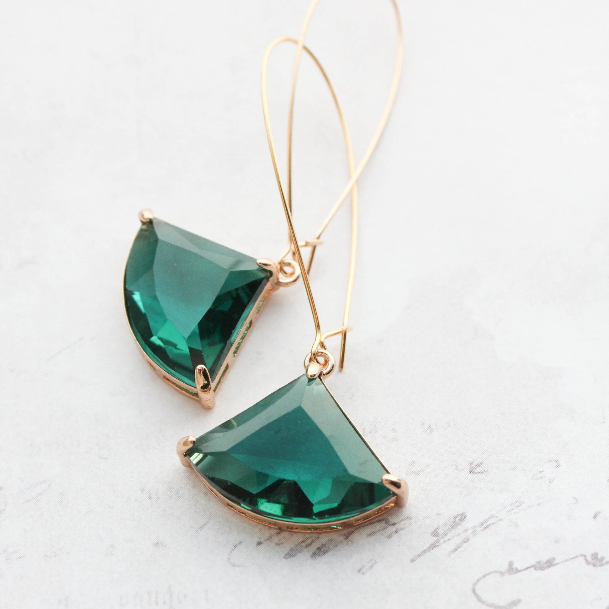 A pair of art deco style emerald green glass earrings with gold prong settings, handing on long ear wires and displayed against a written background.