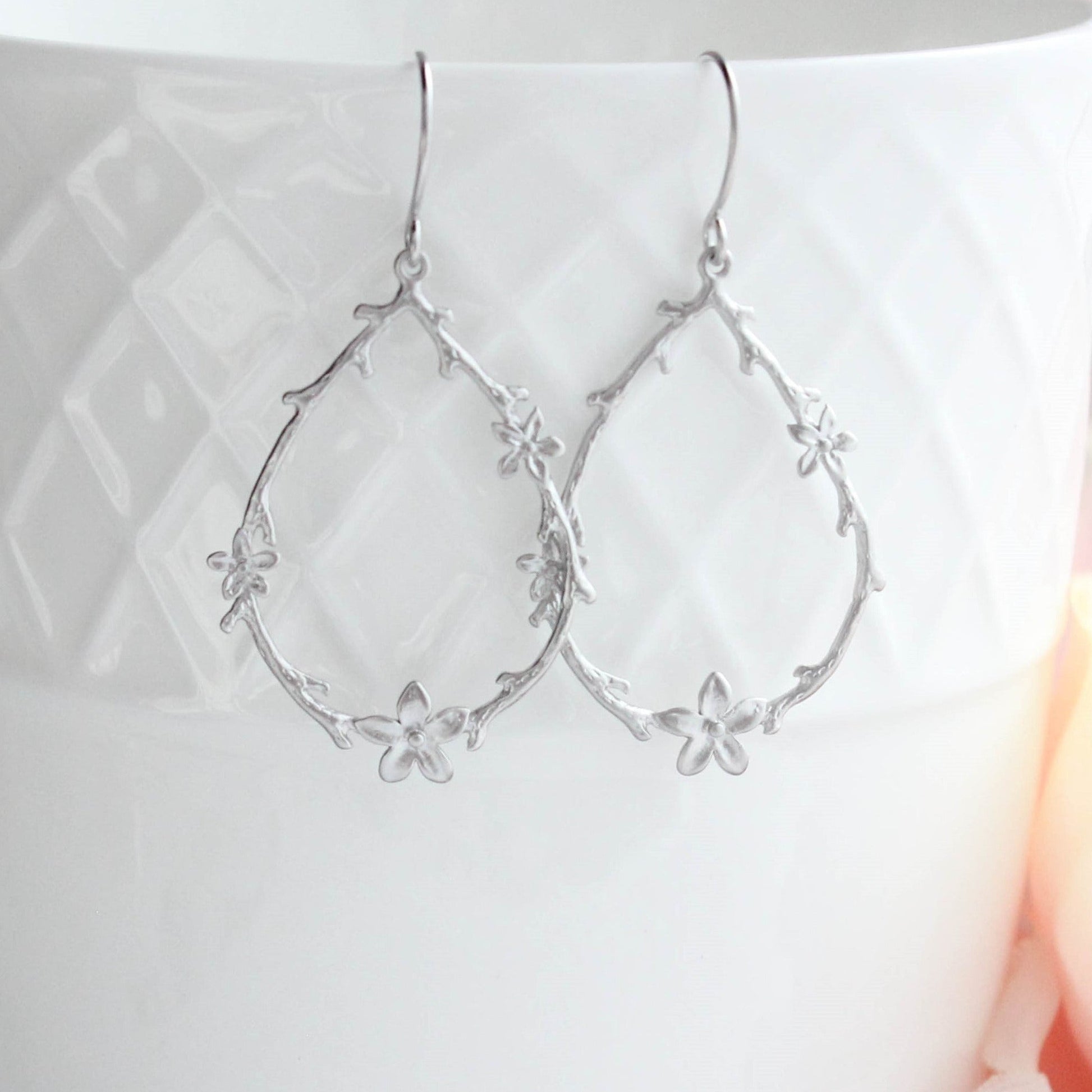 A pair of silver teardrop-shaped dangle earrings with delicate floral and vine details, hanging from a white textured ceramic cup.