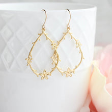 Load image into Gallery viewer, A pair of gold teardrop-shaped hoop earrings with delicate floral vine details, hanging from gold fishhook wires. They are displayed against a white textured ceramic cup with a soft pink rose in the blurred background.
