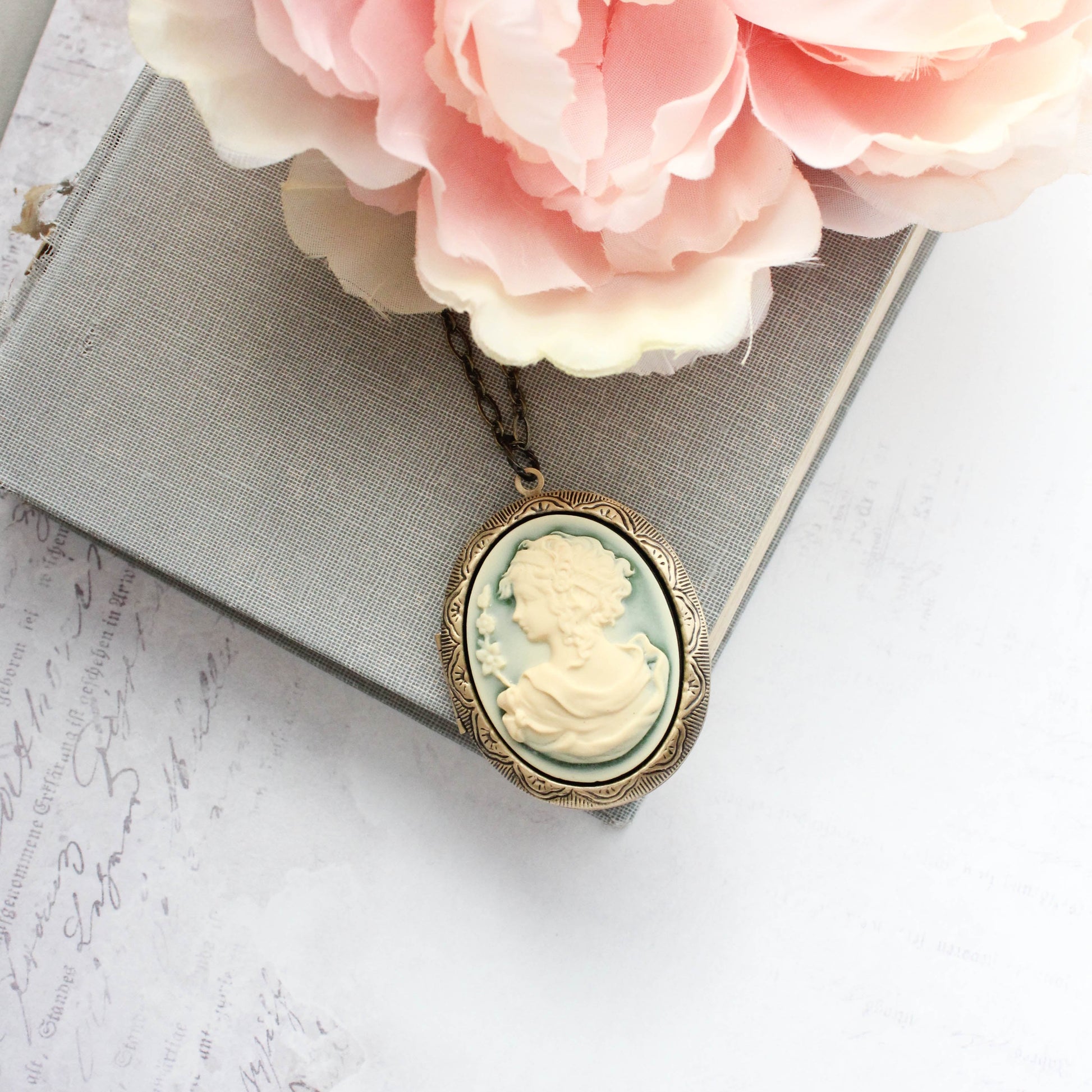 An oval cameo-style pendant necklace with a pale green and cream carved profile design, hanging from a chain and styled on top of a gray book with pink flowers.