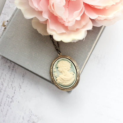 An oval cameo-style pendant necklace with a pale green and cream carved profile design, hanging from a chain and styled on top of a gray book with pink flowers.