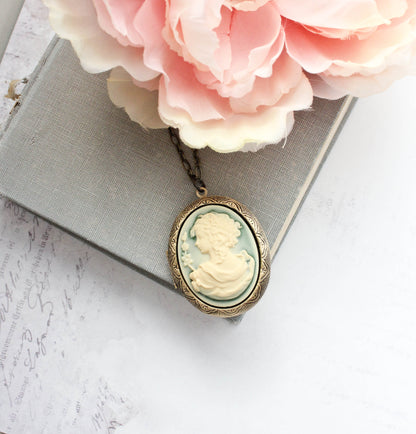 An oval cameo-style pendant necklace with a pale green and cream carved profile design, hanging from a chain and styled on top of a gray book with pink flowers.