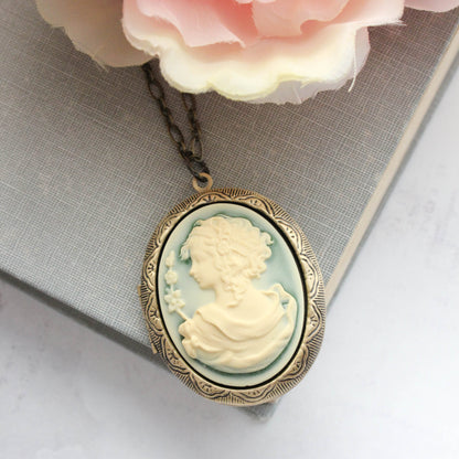 An oval vintage-style cameo pendant necklace with a pale blue-green background and cream-colored profile portrait, set in an antiqued gold-toned frame and hanging on a dark chain. The pendant is styled on a gray textured book beneath soft pink flowers.