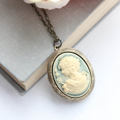 An oval cameo pendant necklace with a pale blue-green background and cream-colored classical bust, hanging from an antique bronze chain. The pendant is styled on top of an old book with pink flowers in the corner.