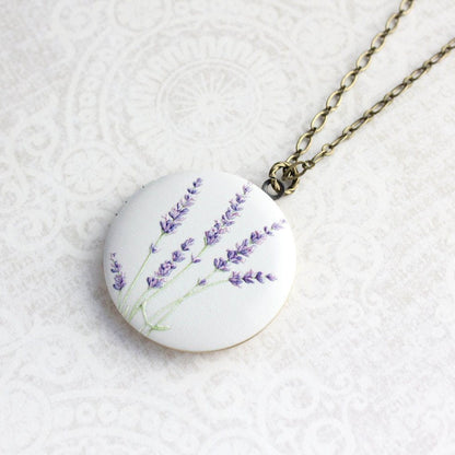 A round white pendant necklace featuring a painted purple lavender bouquet, suspended from an antique bronze chain on a textured white background.