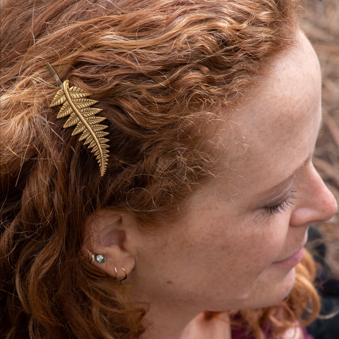 A close-up side profile of a woman with curly red hair wearing a gold fern-shaped hair clip.