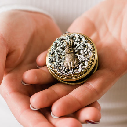 A close-up of a person's hands holding an ornate round locket with a raised rabbit motif and intricate floral detailing. The locket has a warm antique gold finish with silver-toned recessed details, set against a soft, light-colored ribbed fabric background.