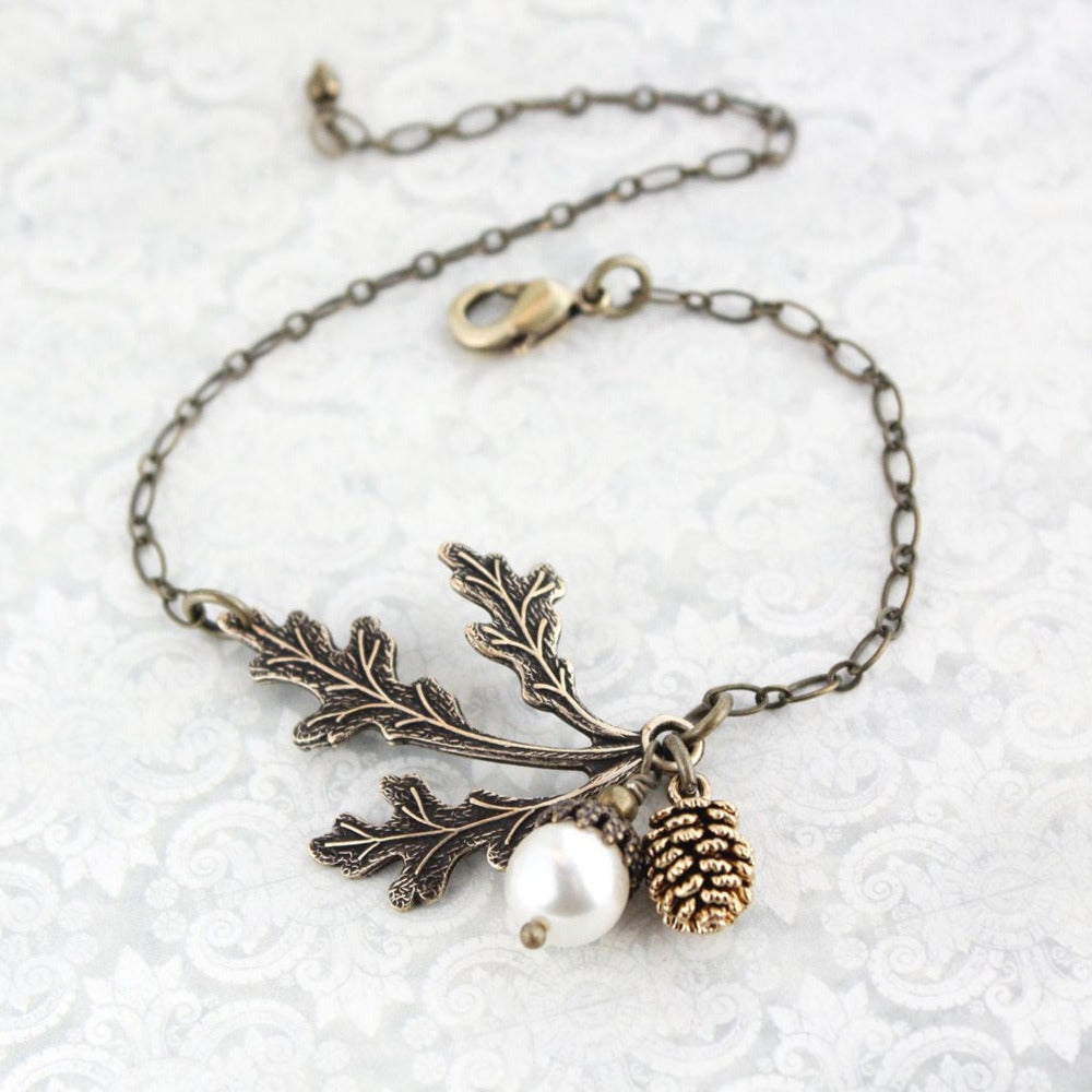 An adjustable bracelet featuring a three leaf branch in antiqued brass, a white pearl acorn, and an antiqued gold pine cone on an antiqued gold chain.
