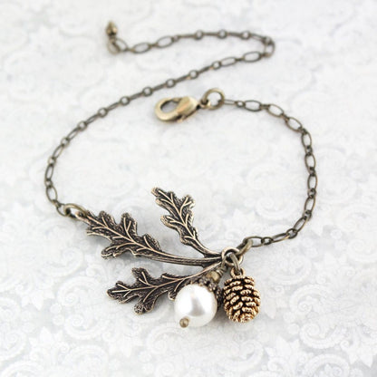 An adjustable bracelet featuring a three leaf branch in antiqued brass, a white pearl acorn, and an antiqued gold pine cone on an antiqued gold chain.