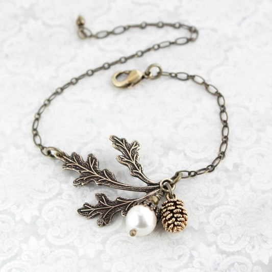 An adjustable bracelet featuring a three leaf branch in antiqued brass, a white pearl acorn, and an antiqued gold pine cone on an antiqued gold chain.
