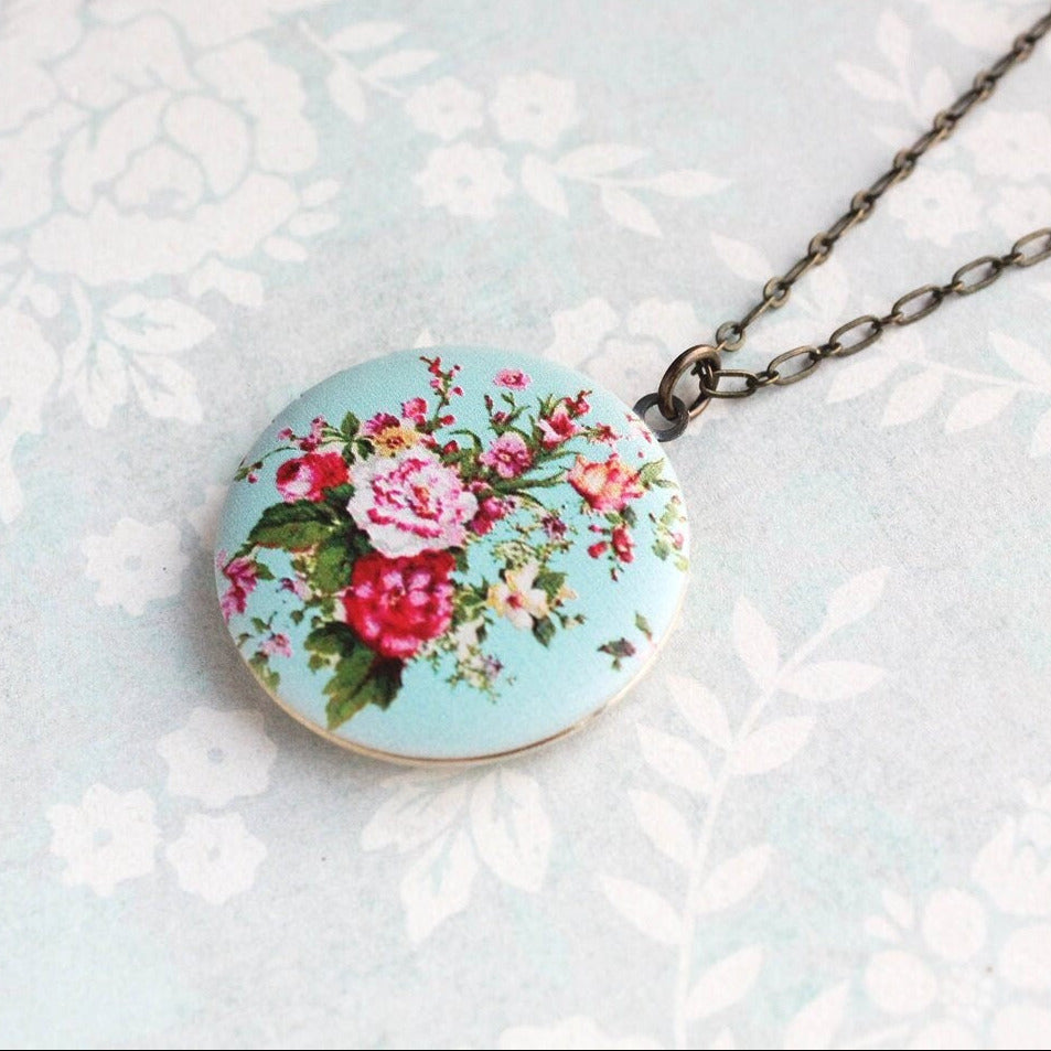 A floral locket necklace with a vintage style motif on an aqua background, attached to an antique brass chain.