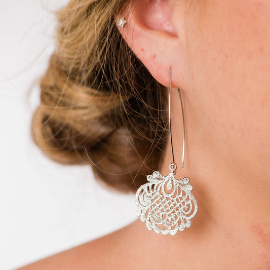 A close-up image of a woman's ear wearing a long silver lace drop dangle earring with a filigree pattern.