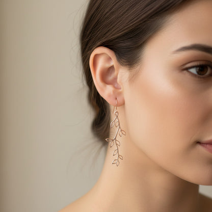 A pair of rose gold branch earrings dangling from a white ceramic cup, with a close-up of the earrings and a pink flower in the background.