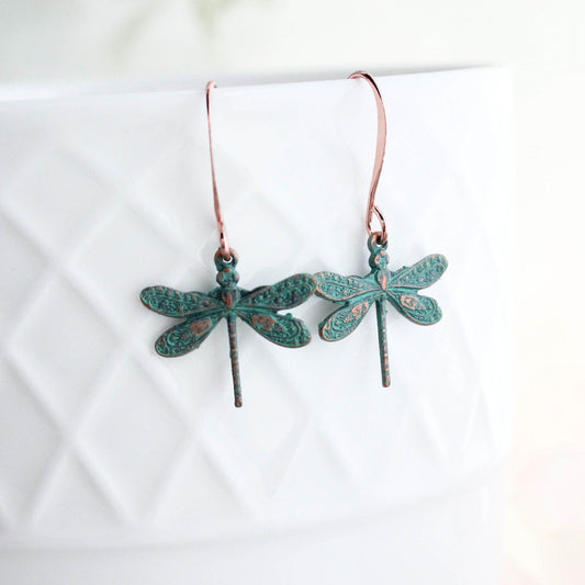 A pair of small dragonfly earrings with a mint patina copper brass finish, displayed on a white surface with a floral background.