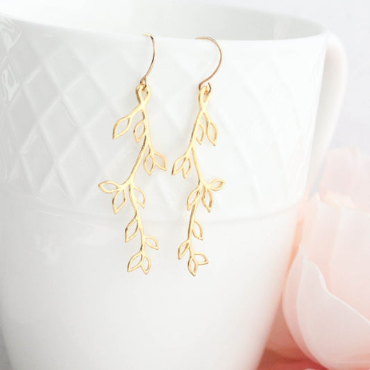 A pair of gold-colored leaf and branch earrings dangling from a white cup