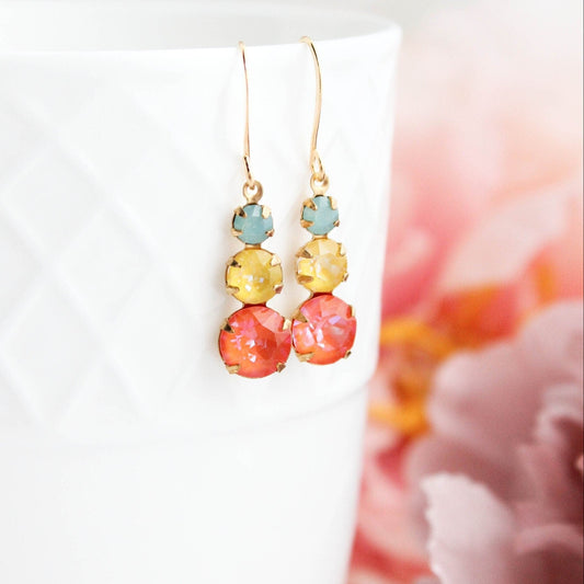 A pair of vintage glass drop earrings in shades of orange and yellow with gold brass rhinestone details, displayed on a white surface with pink flowers in the background.