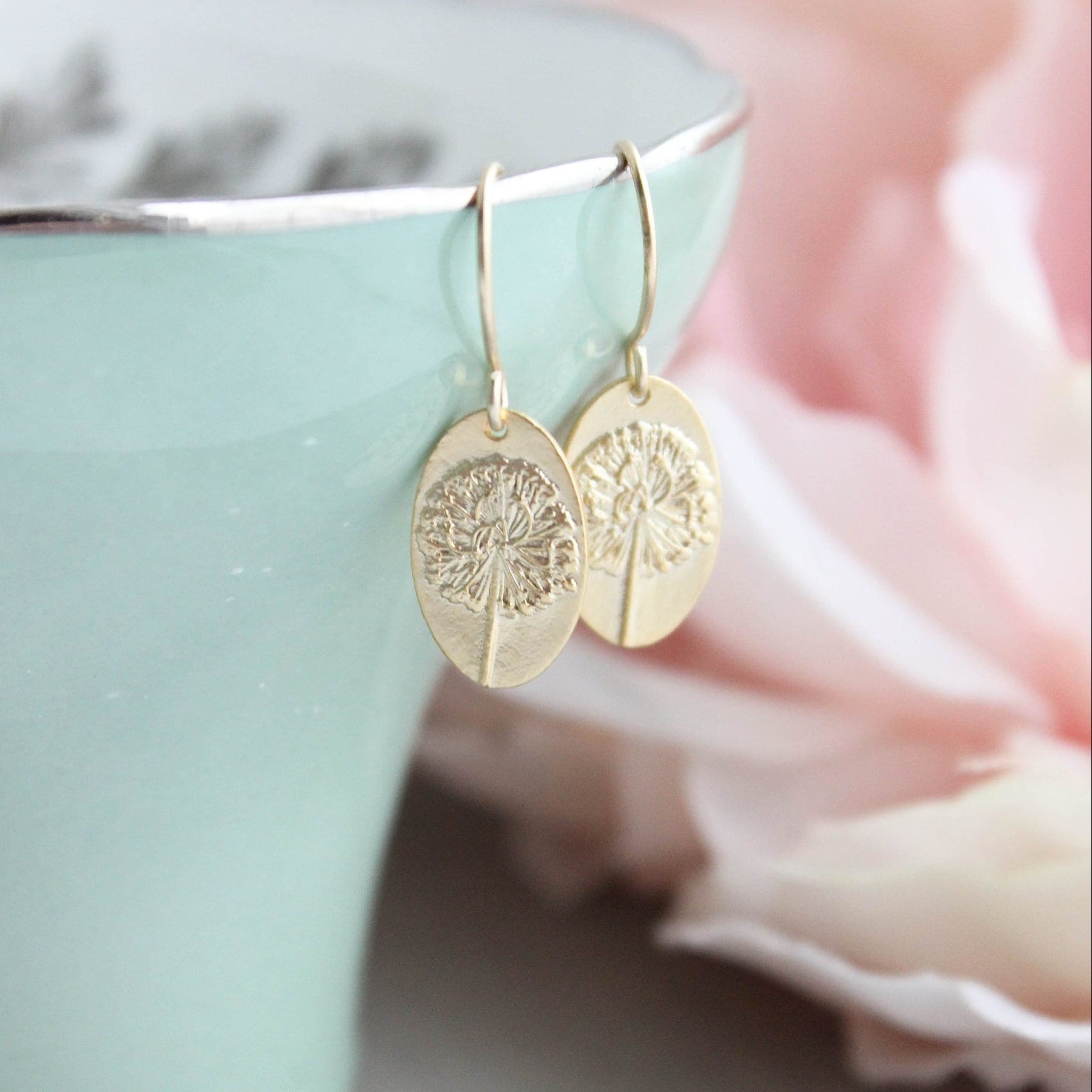 A pair of small oval drop earrings with a raised dandelion flower and stem, gold or silver plated, displayed on a green ceramic cup next to a pink flower.