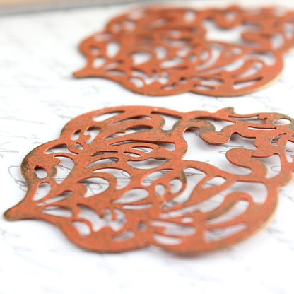 A pair of orange patina filigree dangle earrings with a large drop lace design, placed on a surface with written text.