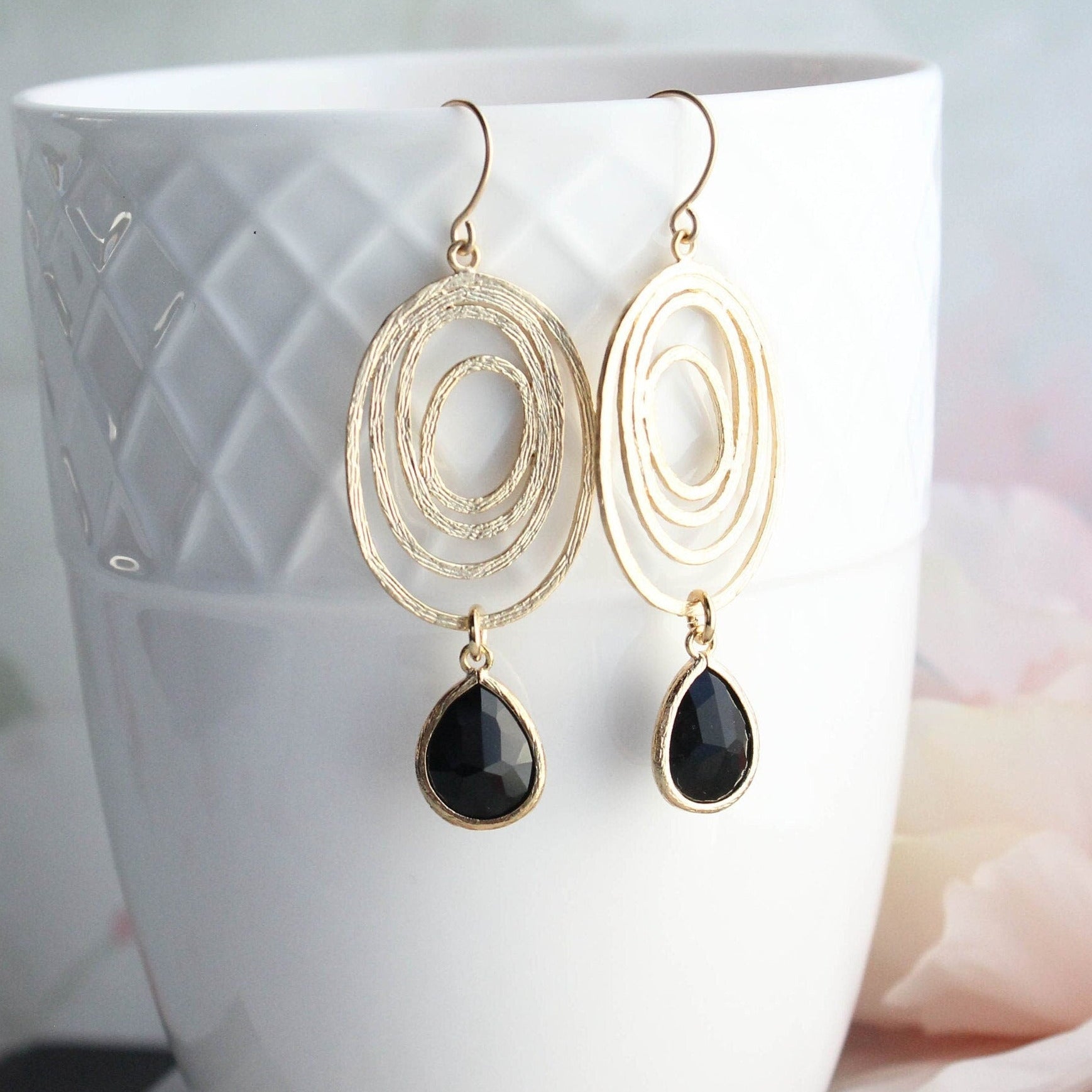 A pair of gold-colored oval swirl earrings with black drop earrings, displayed in a white ceramic cup against a floral background.