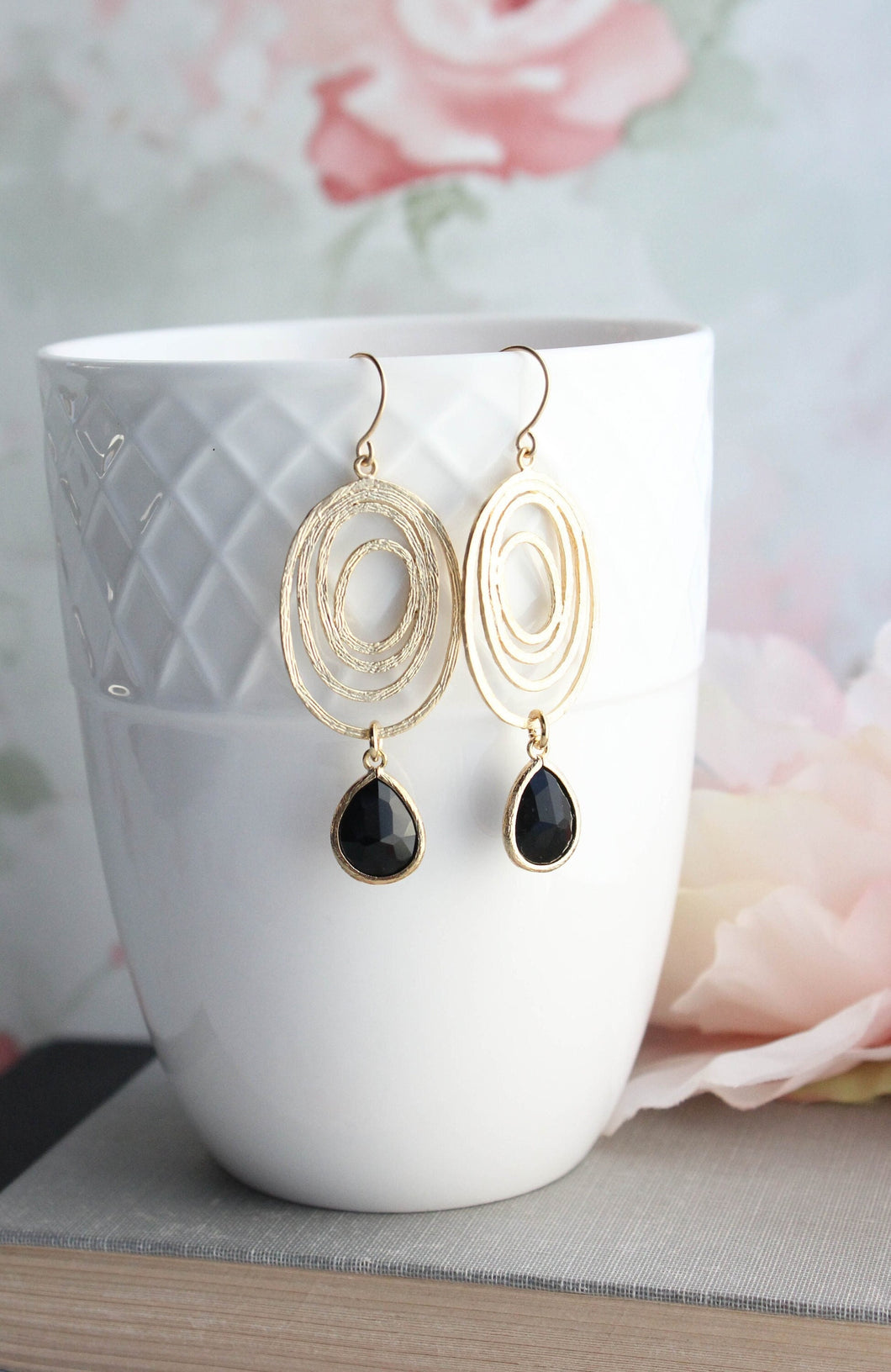 A pair of gold-colored oval swirl earrings with black drop earrings, displayed in a white ceramic cup against a floral background.