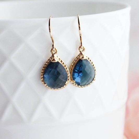 A pair of navy blue glass earrings with gold brass rope settings, displayed on a white earthenware cup with a pink flower in the background.