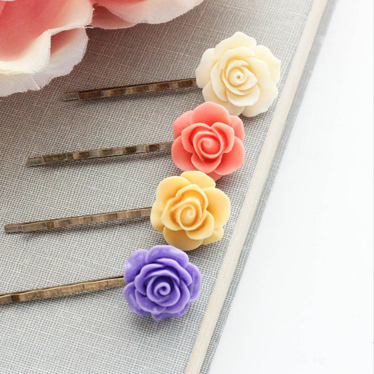A set of four resin rose bobby pins in coral peach, bright violet purple, bright yellow, and ivory cream colors, displayed on a neutral background.