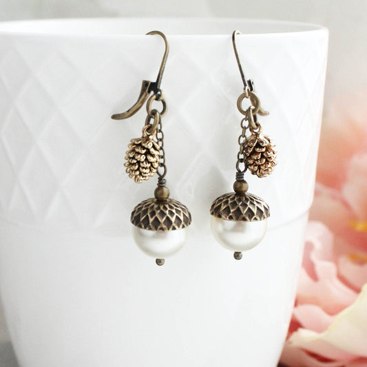 A pair of nature-inspired earrings with pine cone and pearl acorn design, displayed in a white geometric patterned vase with a floral background.