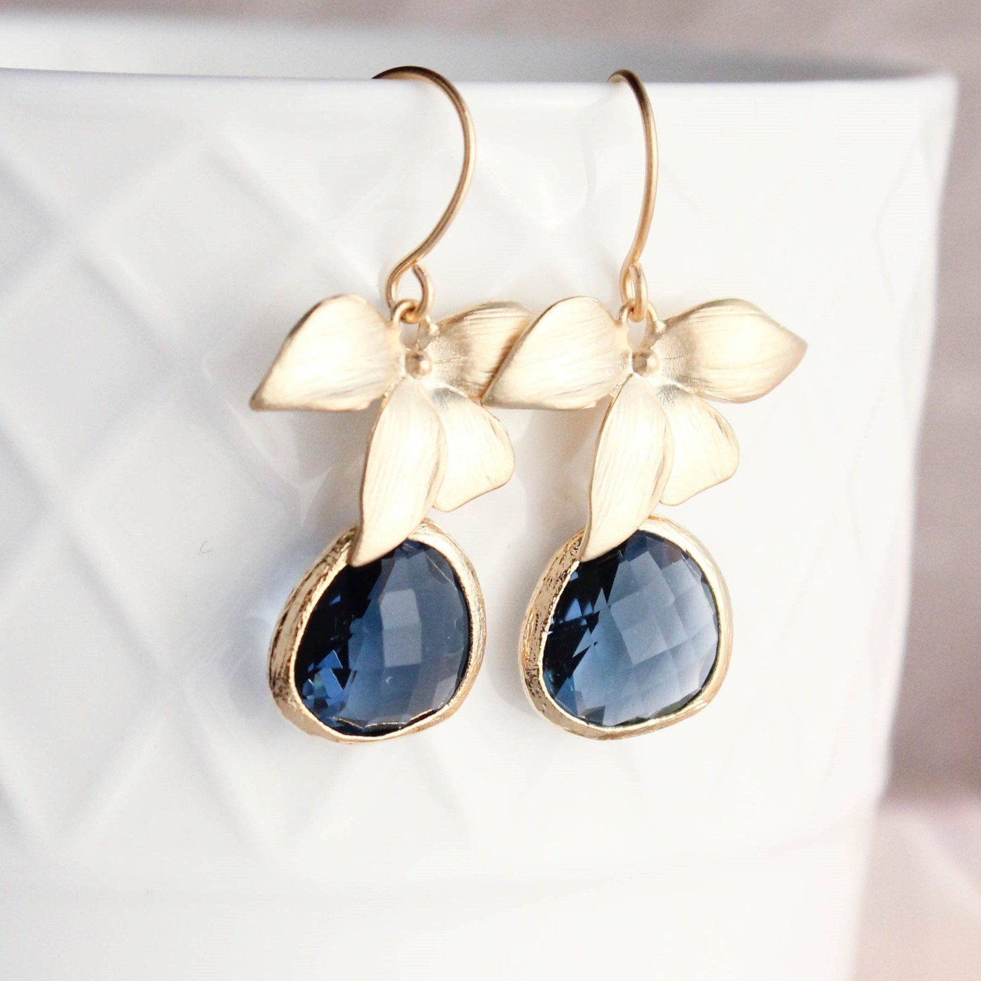 A pair of gold orchid earrings with matte finish and navy blue glass flowers, displayed on a white textured surface.
