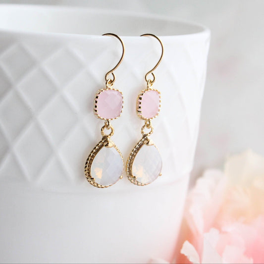 A pair of gold-colored, opal glass dangle earrings with pink and white accents, displayed on a white surface with a floral background.