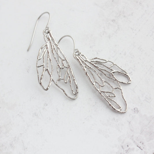 A pair of silver dangling earrings shaped like stylized wings with an open, textured wireframe design, shown on a light gray marble background.