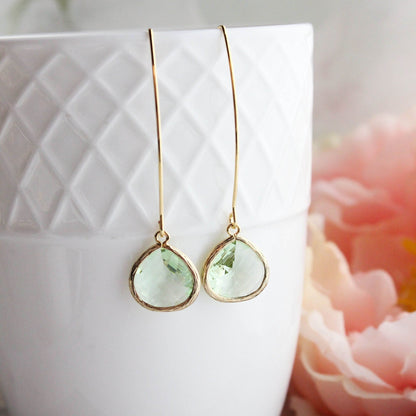 A pair of spring green glass dangle earrings with gold plated brass ear wires, displayed in a white geometric patterned vase with pink flowers in the background.