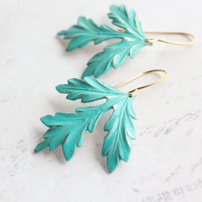 A pair of leaf-shaped earrings with a teal verdigris patina and gold plated brass edges, displayed against a light background.