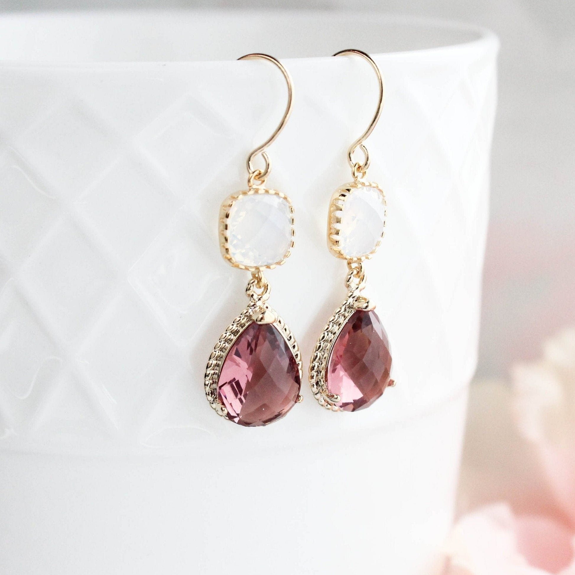 A pair of gold-colored, dusty plum and opal glass dangle earrings with gold rope settings, displayed on a white surface with pink accents.
