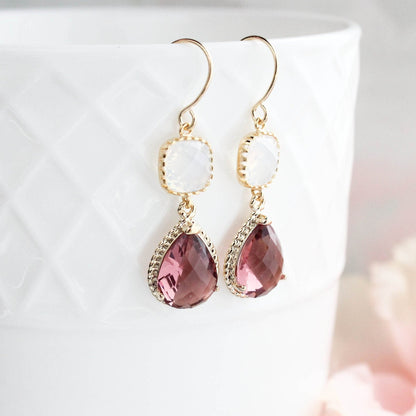 A pair of gold-colored, dusty plum and opal glass dangle earrings with gold rope settings, displayed on a white surface with pink accents.