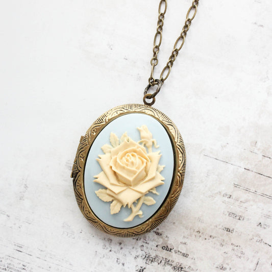 An oval locket necklace with a vintage brass frame and a light blue cream rose resin cameo centerpiece, displayed on a textured white background.