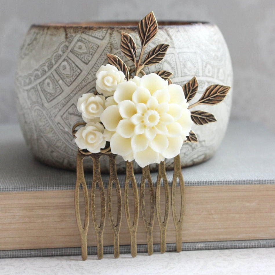 A cream-colored floral hair comb with roses and big dahlias, set on antique brass filigree with gold brass leaves, displayed on a textured surface.