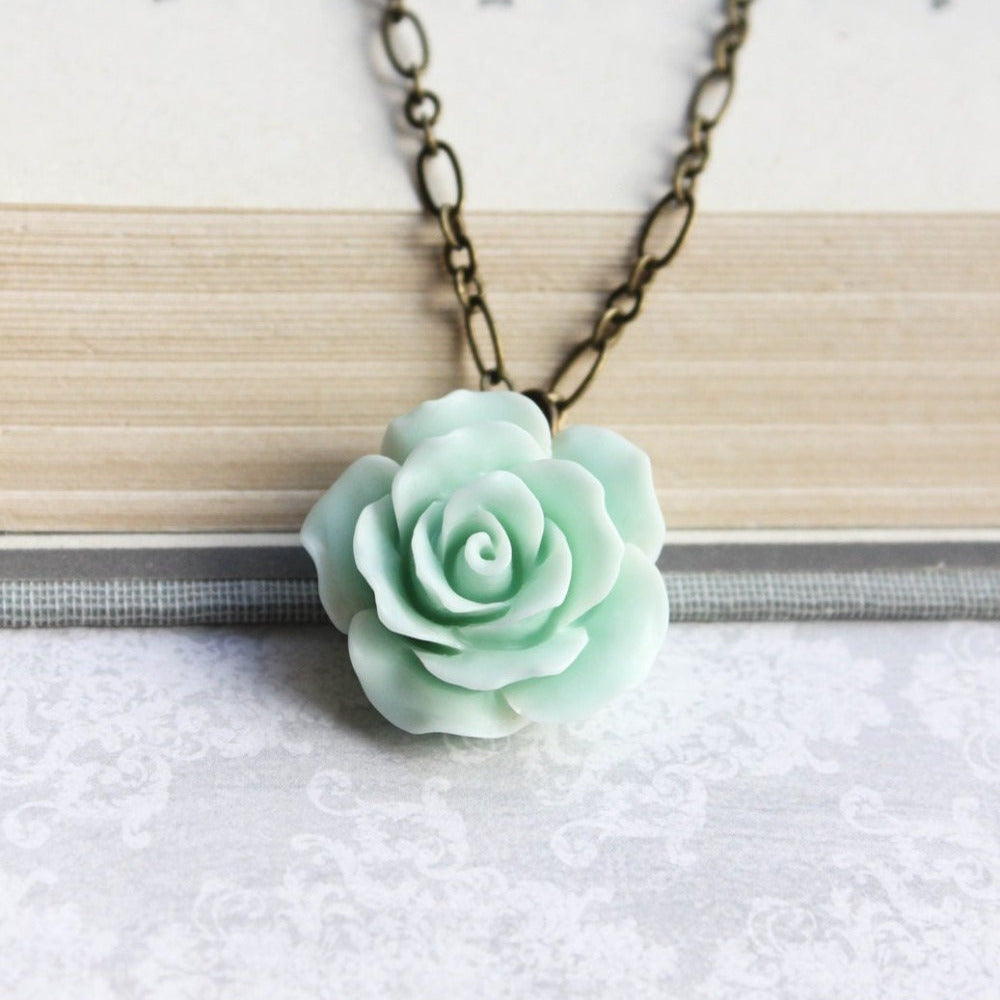 A seafoam mint green floral pendant necklace with a ruffled petal rose design, attached to a 20-inch antique brass chain, displayed on a book.