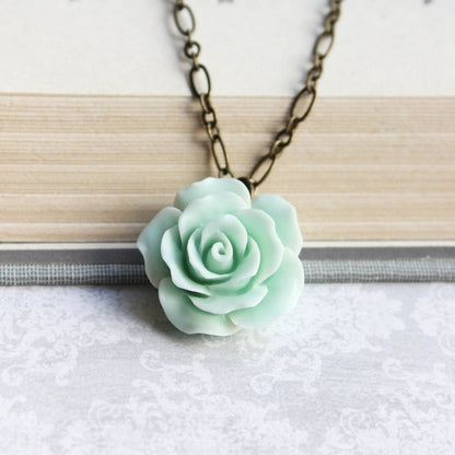 A seafoam mint green floral pendant necklace with a ruffled petal rose design, attached to a 20-inch antique brass chain, displayed on a book.