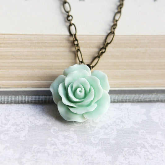 A seafoam mint green floral pendant necklace with a ruffled petal rose design, attached to a 20-inch antique brass chain, displayed on a book.