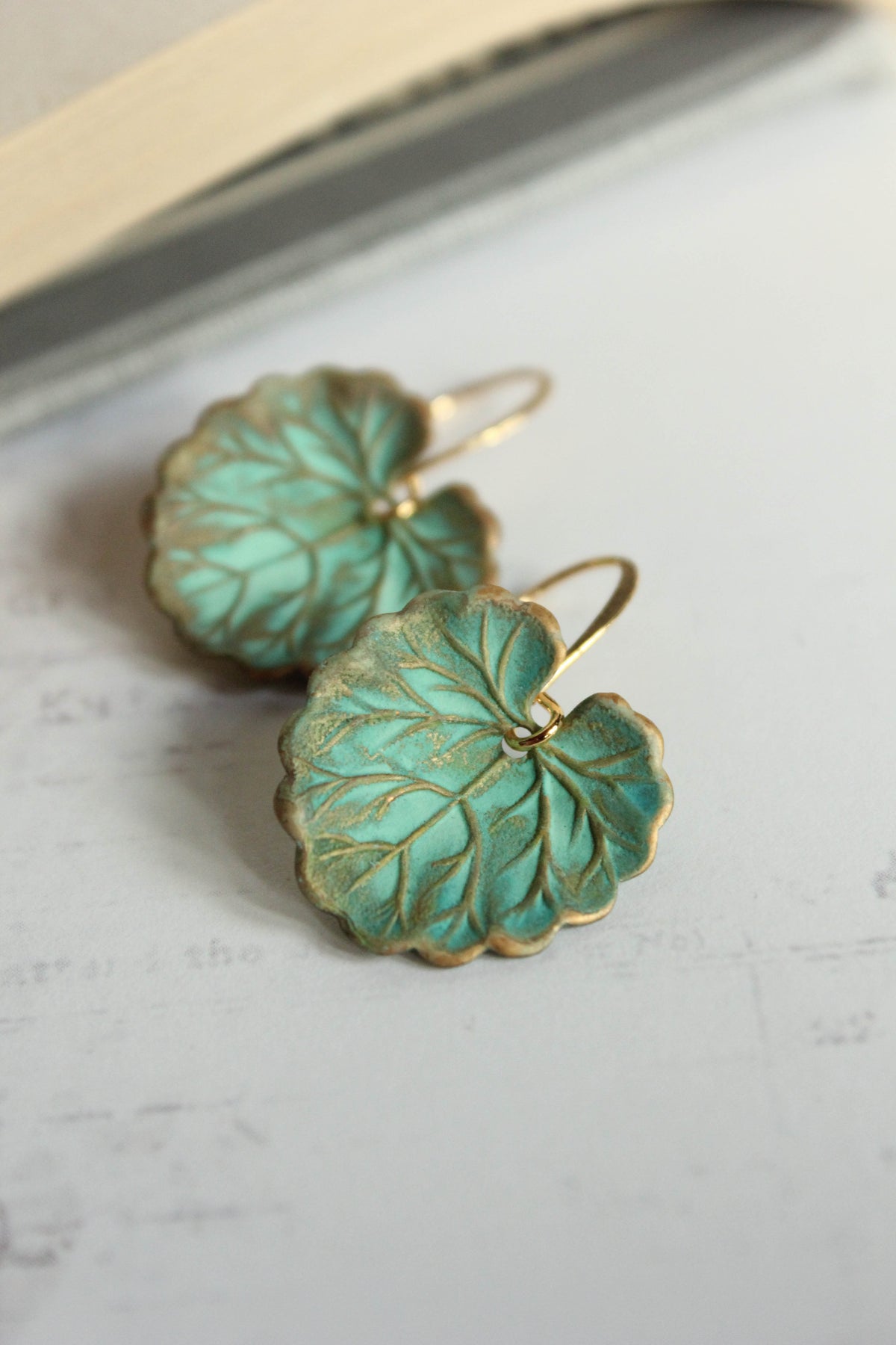 A pair of green leaf earrings with a rustic verdigris patina finish placed on a white surface.