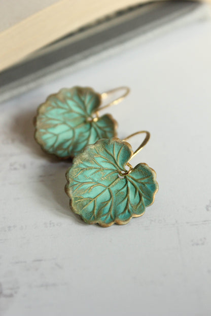 A pair of green leaf earrings with a rustic verdigris patina finish placed on a white surface.