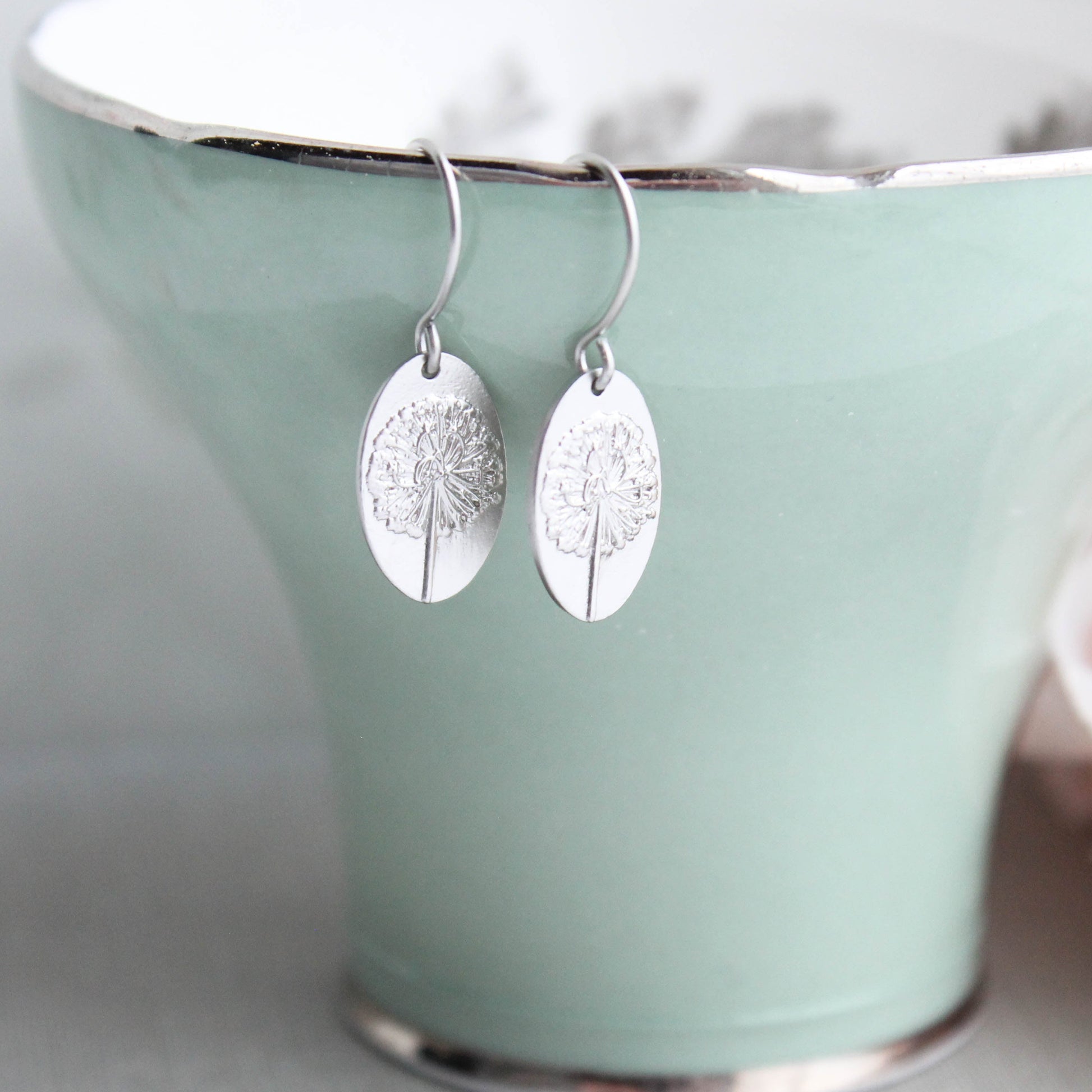 A pair of matte silver-plated oval drop earrings with a raised dandelion flower and stem, displayed on a green ceramic cup.