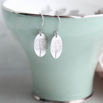 A pair of matte silver-plated oval drop earrings with a raised dandelion flower and stem, displayed on a green ceramic cup.