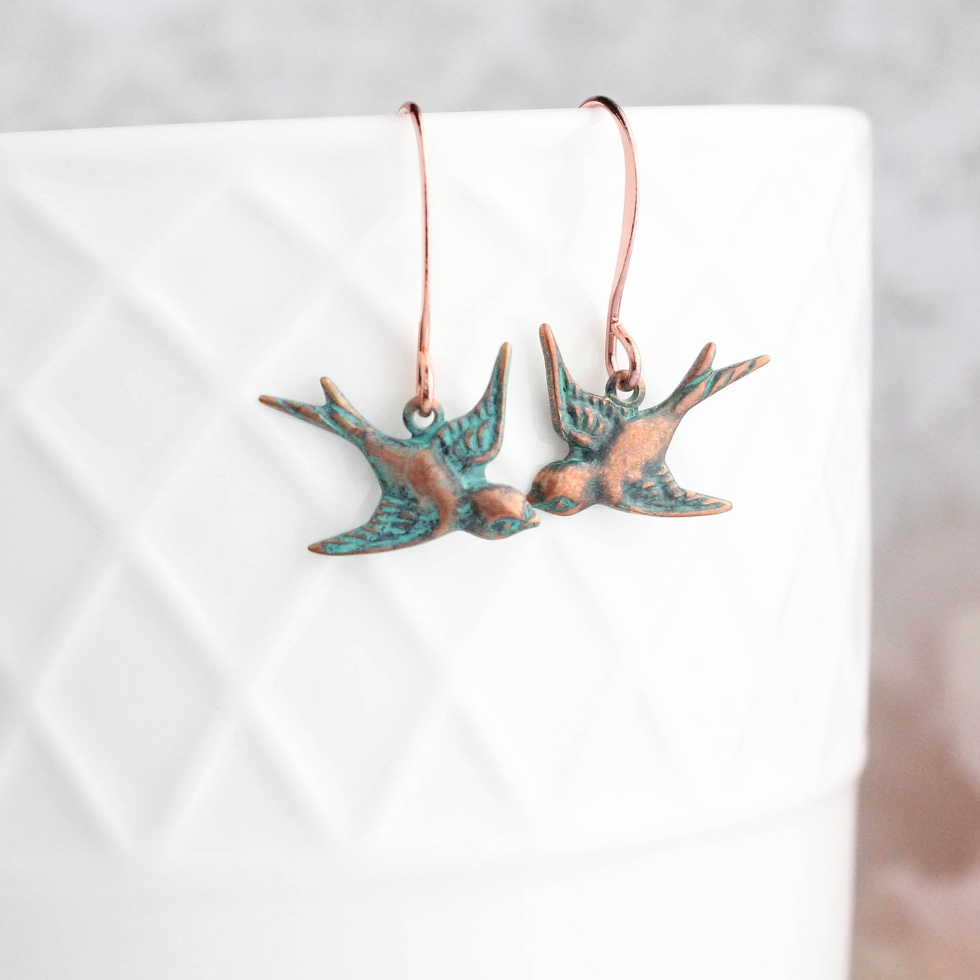 A pair of copper patina small flying bird earrings displayed on a white earthenware surface.