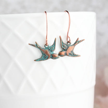 A pair of copper patina small flying bird earrings displayed on a white earthenware surface.