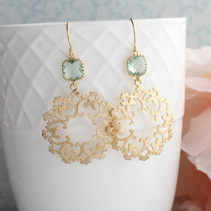 Gold filigree dangle earrings with aqua glass stones, displayed in a white vase with a lace filigree design at the bottom.