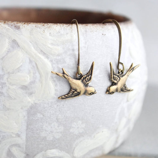A pair of gold brass earrings designed as small flying birds, displayed on a textured surface.