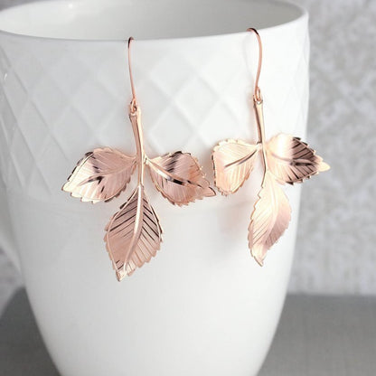 A pair of rose gold leaf branch earrings dangling from a white geometric patterned cup.