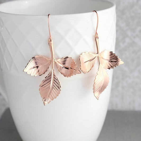 A pair of rose gold leaf branch earrings dangling from a white geometric patterned cup.